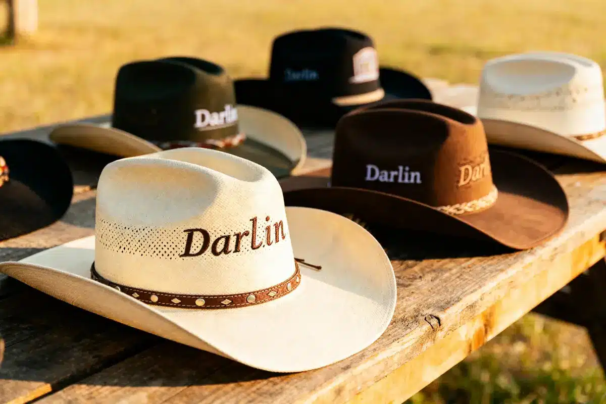 Various cowboy hats on wooden table, each embroidered with "Darlin," set against a grassy outdoor background.