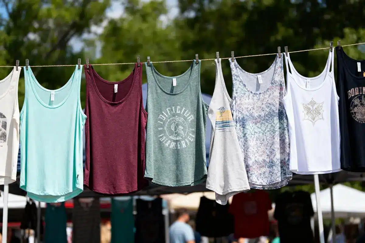 Colorful tank tops hanging on a clothesline at an outdoor market, with trees and tents in the background.