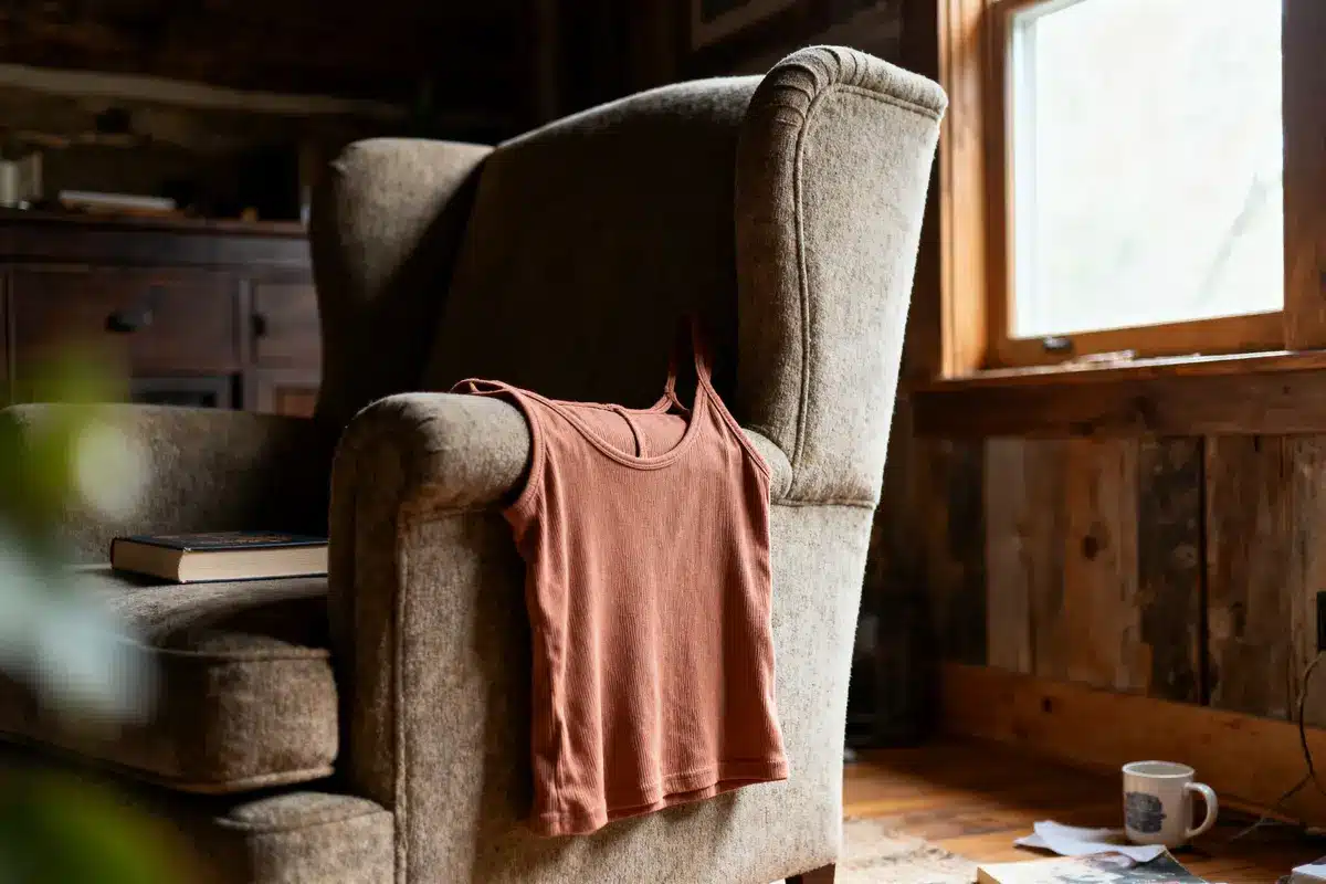 Brown tank top on armchair, with book on seat; window, mug, and wooden walls in cozy, rustic room.