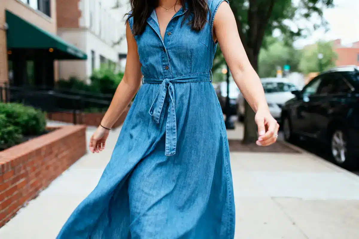 Woman in a blue denim dress walking on a city sidewalk, surrounded by trees and buildings.