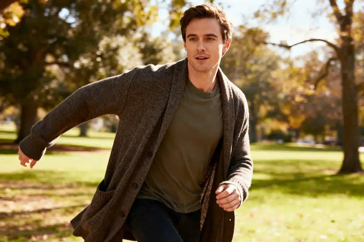 Man in cozy cardigan running outdoors in sunny park with autumn trees in the background.