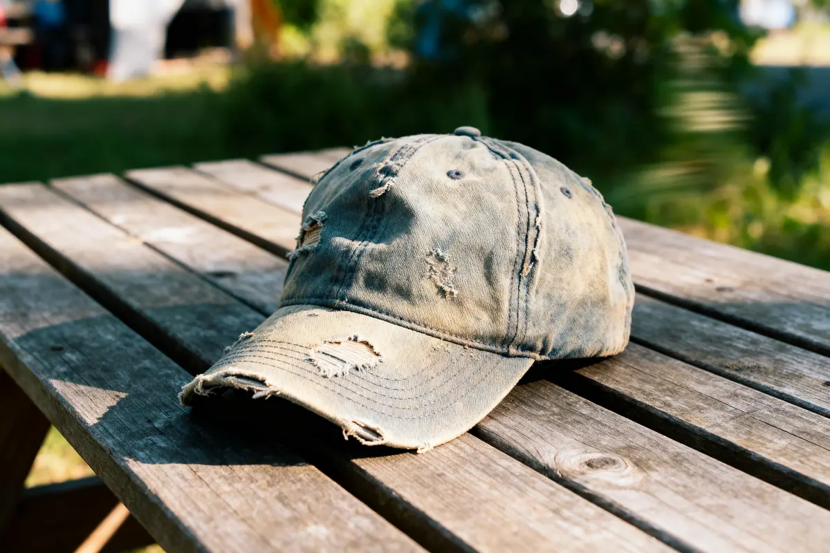 A worn, distressed baseball cap on a wooden picnic table under sunlight, with blurred greenery in the background.