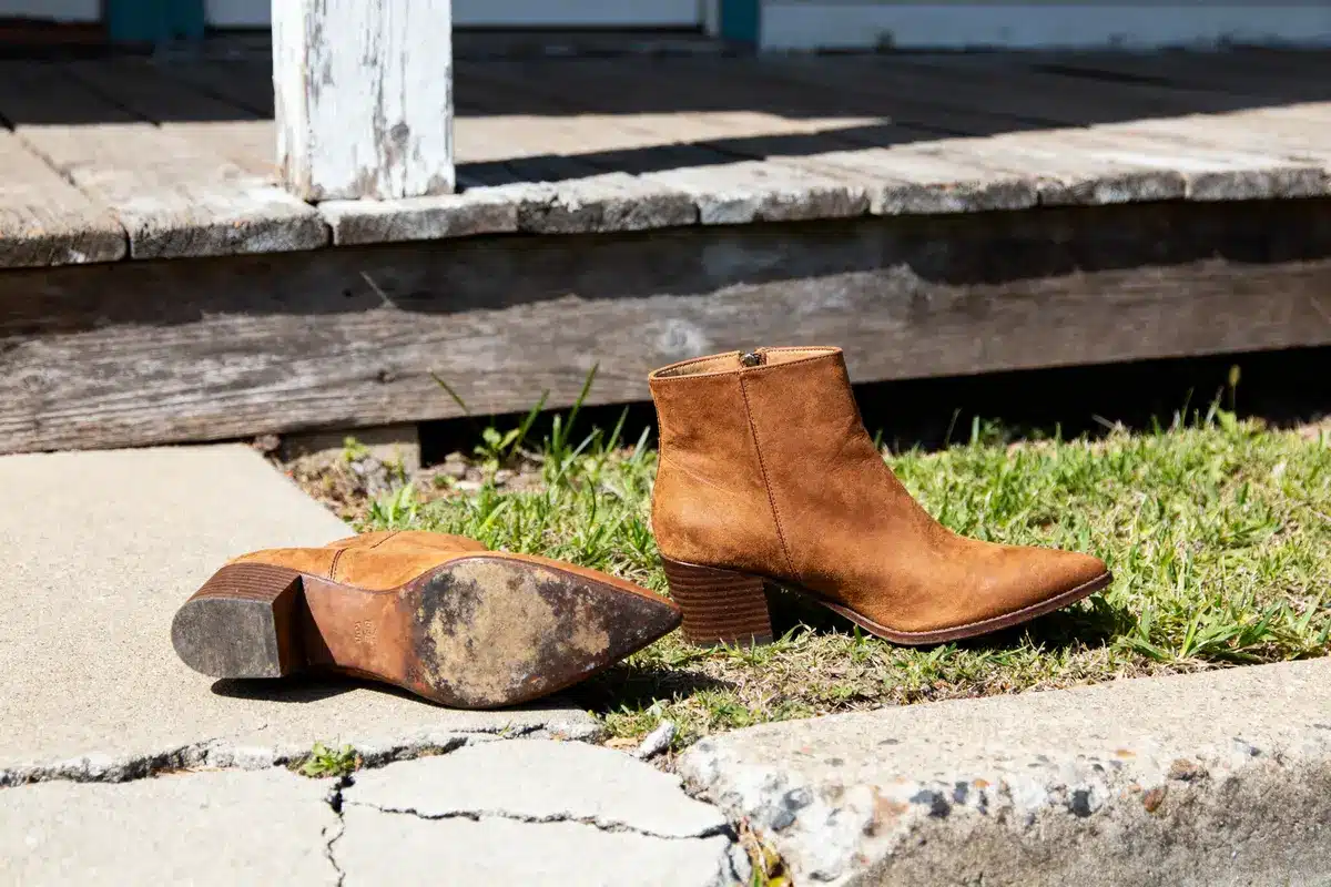 Brown suede ankle boots on grass near wooden porch and cracked concrete pathway.