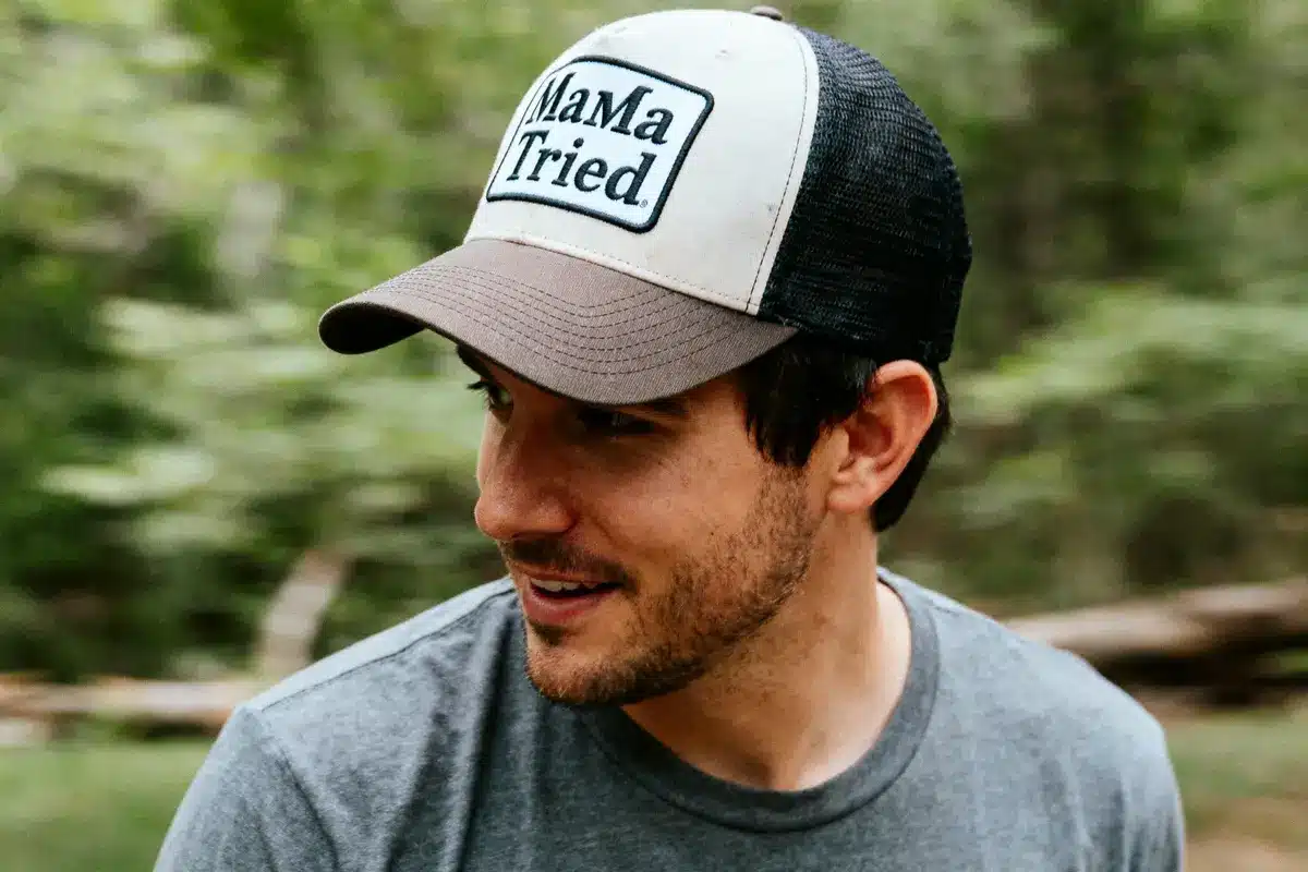 Man wearing a "Mama Tried" cap and gray T-shirt, looking sideways in a forest setting.