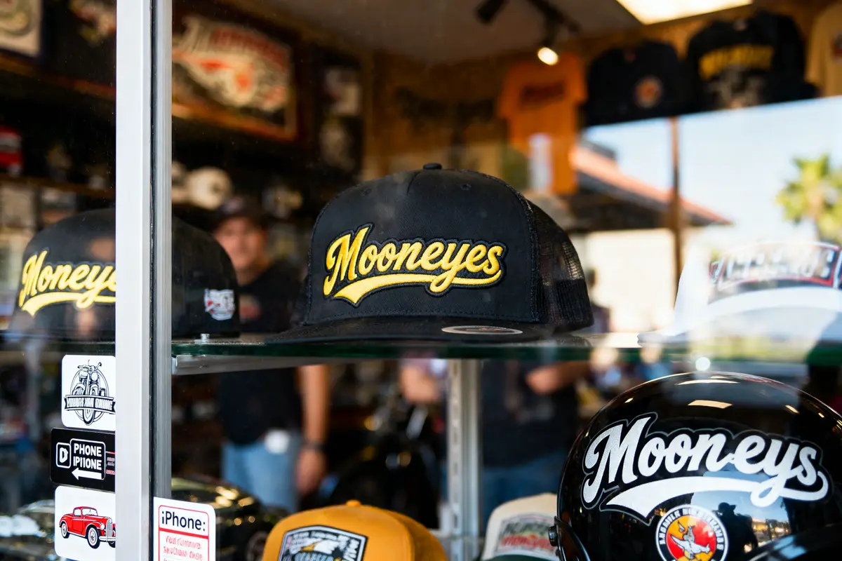 Black "Mooneyes" cap and helmet on display in a shop with branding and stickers in the background.