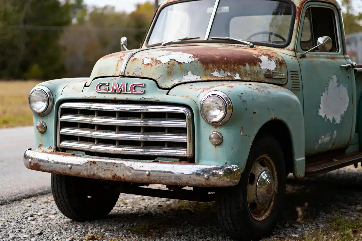 Rusty vintage GMC truck with peeling paint, chrome grill, parked on gravel road, surrounded by blurred trees.