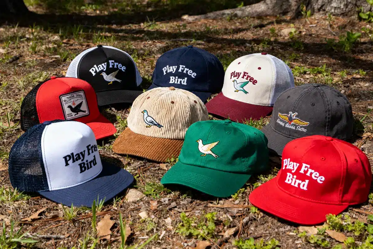 Collection of colorful baseball caps on grass, featuring bird designs and "Play Free Bird" text embroidery.